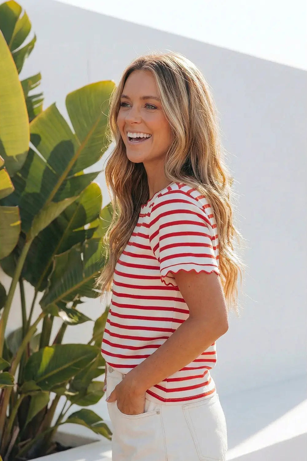 Woman wearing a red and white striped shirt standing next to a plant against a white wall.