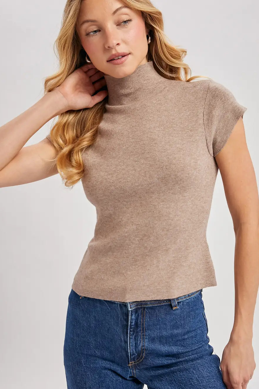 A woman wearing a fitted short-sleeve mock neck top in a heathered taupe-brown color, paired with blue jeans, against a light gray studio background.