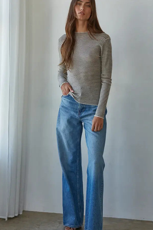 A woman modeling a beige and gray striped long-sleeve knit top with wide-leg blue jeans, standing in a bright room with white walls and a curtain.