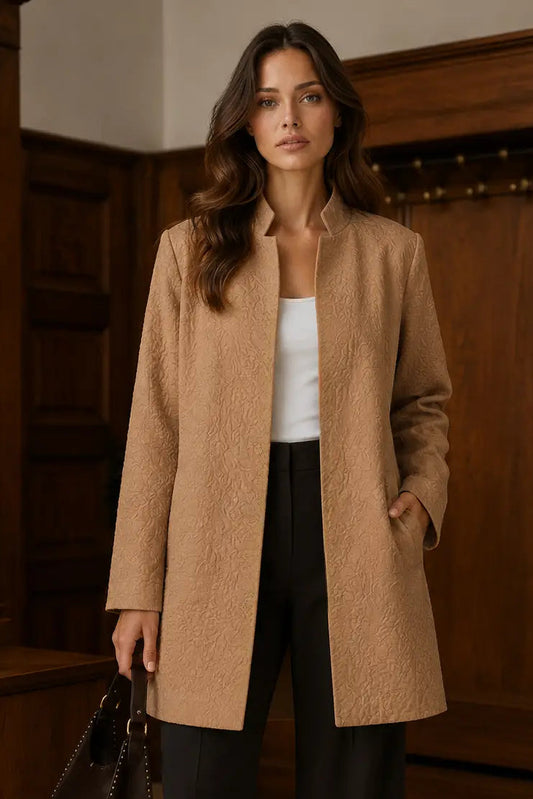 A woman stands indoors wearing a camel-colored textured coat over a white top and black pants, holding a dark handbag. The background shows warm wood paneling and a coat rack.