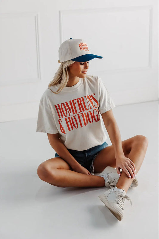 Person wearing a 'Home Runs & Hotdogs' t-shirt and cap sitting on a white floor.