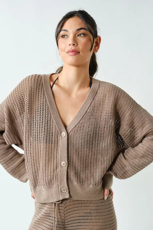 A woman modeling a taupe brown open-knit cardigan sweater with a V-neck and button closure, paired with a matching knit skirt, against a light gray studio background.
