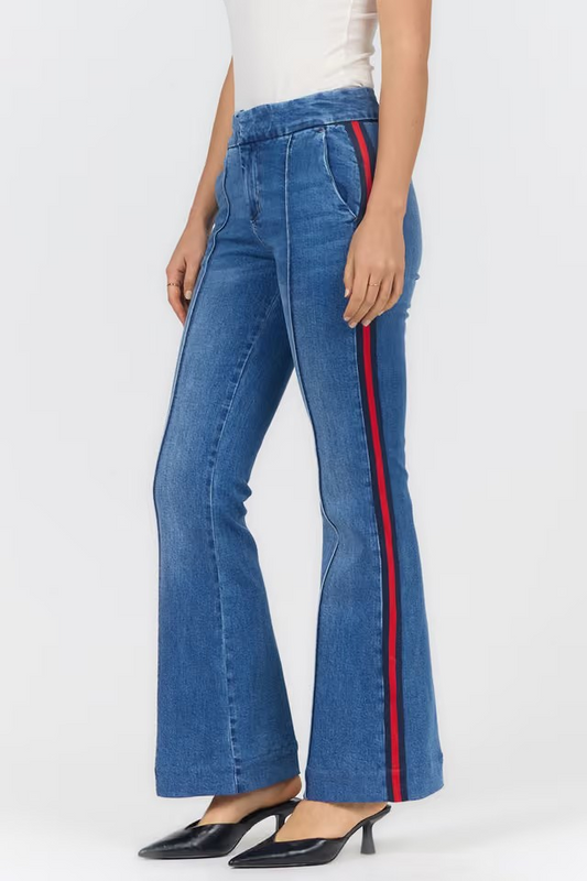 A model wearing blue denim flare jeans with a red and black side stripe, paired with a white top and black pointed-toe mule heels, shown from the waist down on a light gray studio background.