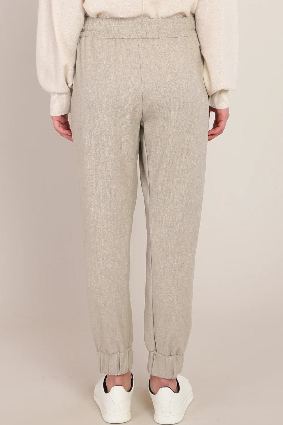 A person shown from the back wearing light beige jogger-style pants with an elastic waistband and cuffed ankles, paired with a cream sweater and white sneakers against a plain beige studio background.
