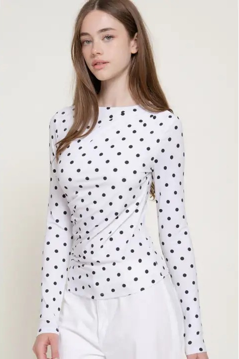 A woman modeling a white long-sleeve top with a black polka dot pattern, paired with white pants, against a plain light beige background.