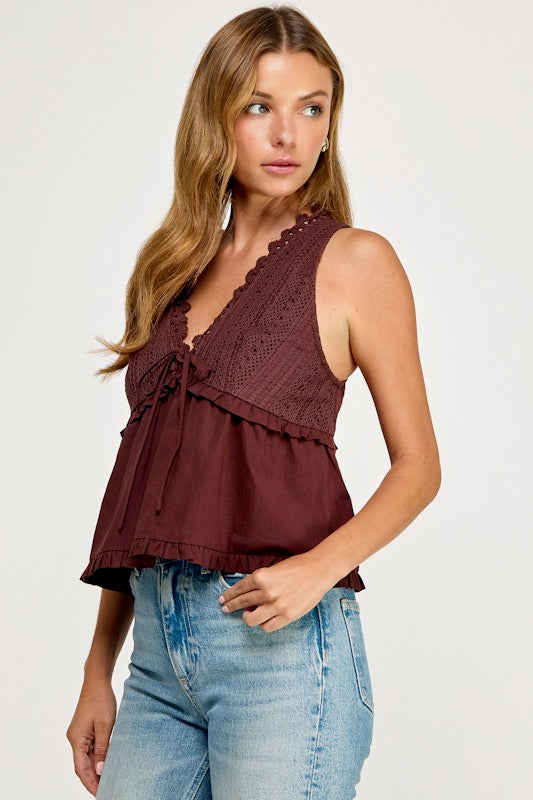 A woman modeling a sleeveless burgundy lace and mesh top with ruffles, paired with light blue jeans, against a plain light gray background.