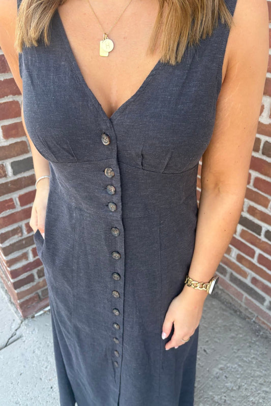 Woman wearing a sleeveless denim dress with buttons against a brick wall.
