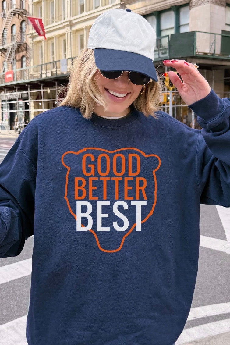 A woman wearing a navy blue sweatshirt with a 'GOOD BETTER BEST' graphic in orange and white, styled with a white and navy baseball cap and sunglasses, standing on a city street crosswalk.