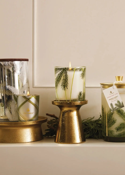 A styled holiday candle display on a shelf, featuring a central white pillar candle with green pine needles inside a clear glass cylinder, set in a gold metal holder. Flanking it are other decorative candles and glass containers with gold and silver accents, pine needles, and a hanging product tag.