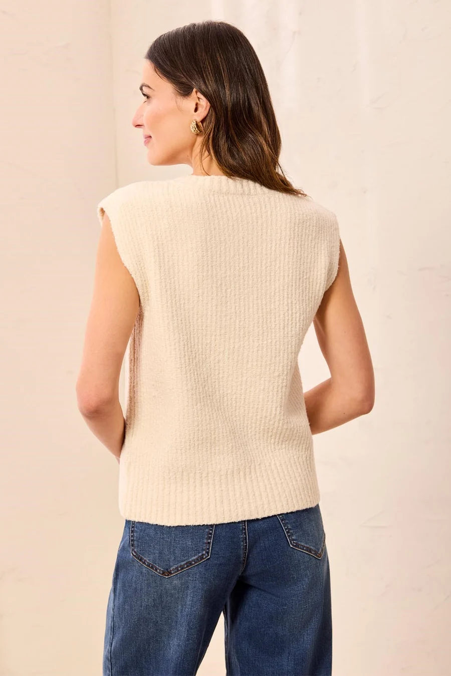 A woman shown from the back wearing a ribbed knit sleeveless vest in cream, paired with blue jeans, against a light beige textured wall.