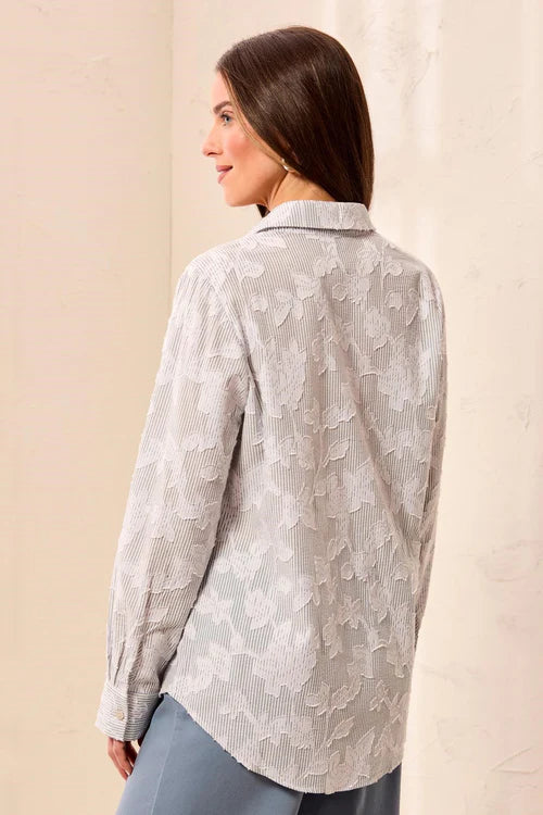 A woman shown from the back wearing a light gray and white textured floral lace blouse, paired with blue-gray pants, against a neutral beige background.