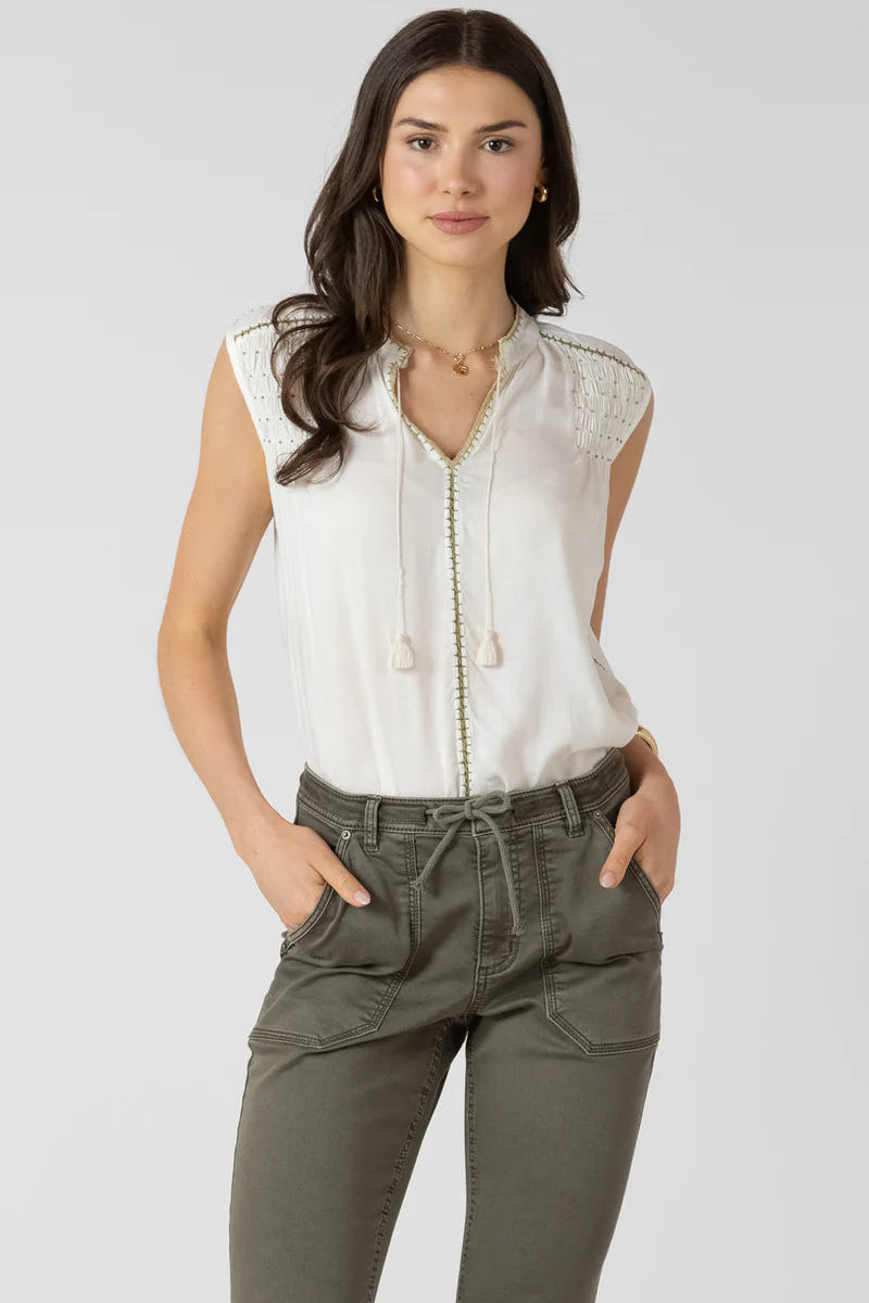 Woman wearing a white sleeveless blouse with lace details and olive green pants on a plain background