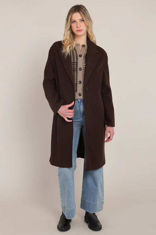 A woman modeling a long dark brown wool-blend coat over a beige plaid sweater, light blue wide-leg jeans, and black boots, photographed against a plain beige studio background.