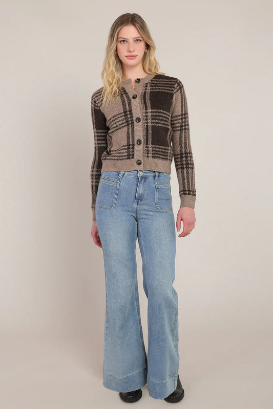 A woman modeling a brown and beige plaid button-up cardigan sweater paired with light-wash blue flare jeans, standing against a neutral beige studio background.