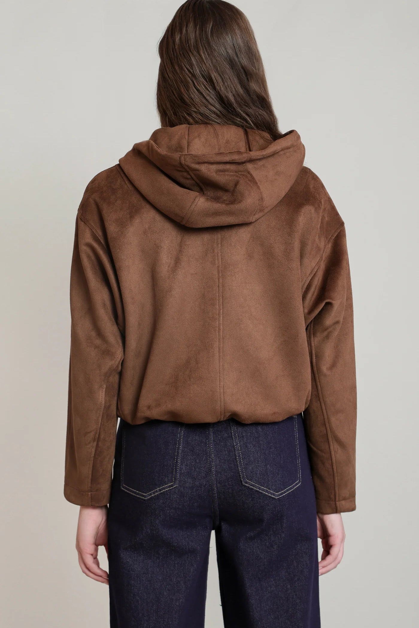A person shown from the back wearing a brown hooded jacket with a suede-like finish and dark blue jeans, against a plain light gray studio background.