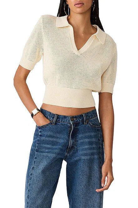 A woman wearing a cream-colored, short-sleeve knit polo-style top with a V-neck and textured openwork pattern, paired with blue jeans, against a plain white background.