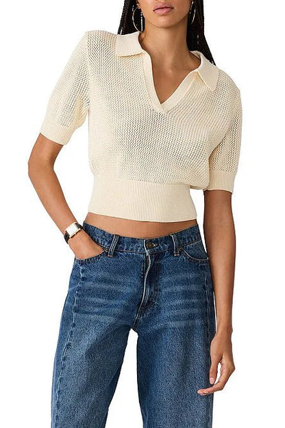 A woman wearing a cream-colored, short-sleeve knit polo-style top with a V-neck and textured openwork pattern, paired with blue jeans, against a plain white background.