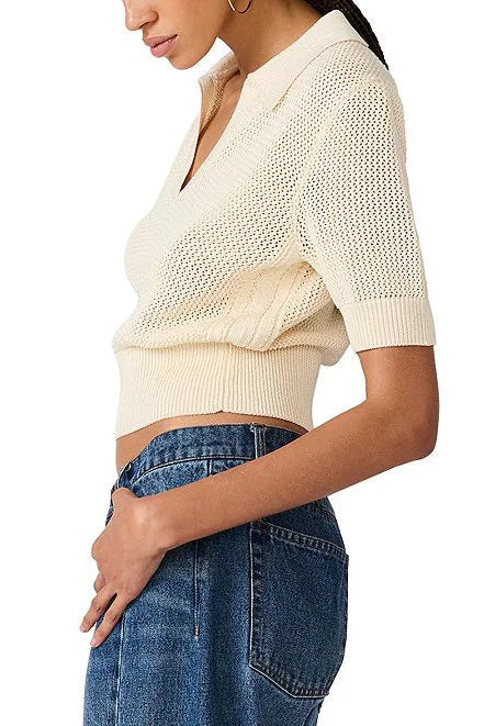 A woman wearing a cream-colored short-sleeve knit polo crop top and blue jeans, shown from a side profile against a plain white background.