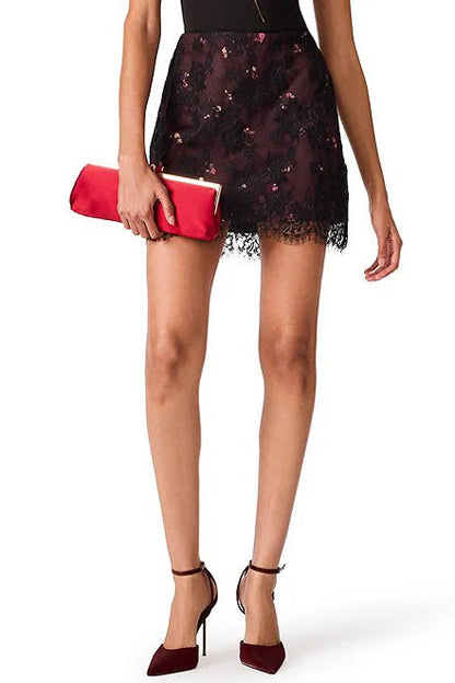 A model wearing a short black lace mini skirt with a red floral pattern, holding a red clutch, and wearing burgundy high heels against a white background.