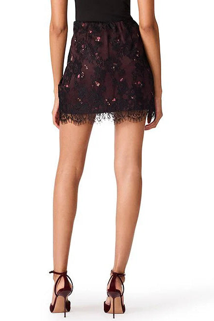 A model shown from the back wearing a short black lace mini skirt with floral embroidery and a scalloped lace hem, paired with dark brown strappy high heels against a white background.