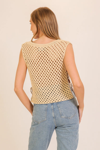 A woman shown from the back wearing a sleeveless beige crochet knit top and light blue jeans against a plain cream background.