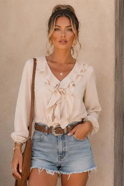 A woman wearing a cream ruffled long-sleeve blouse tucked into light-wash denim shorts with a brown belt, standing against a textured beige wall.