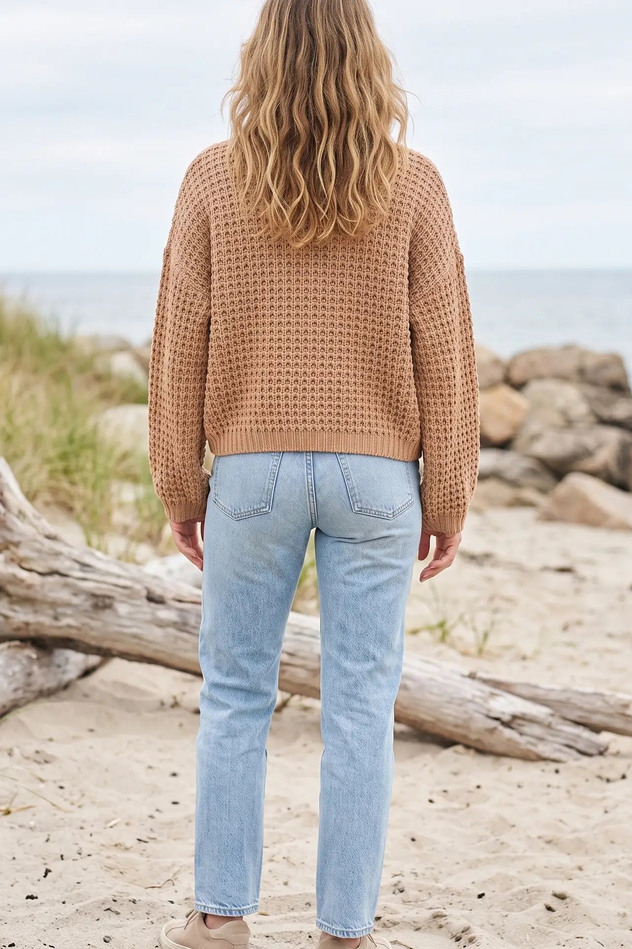 A woman viewed from behind wearing a tan open-knit sweater and light blue jeans, standing on a sandy beach with driftwood and rocks in the background.