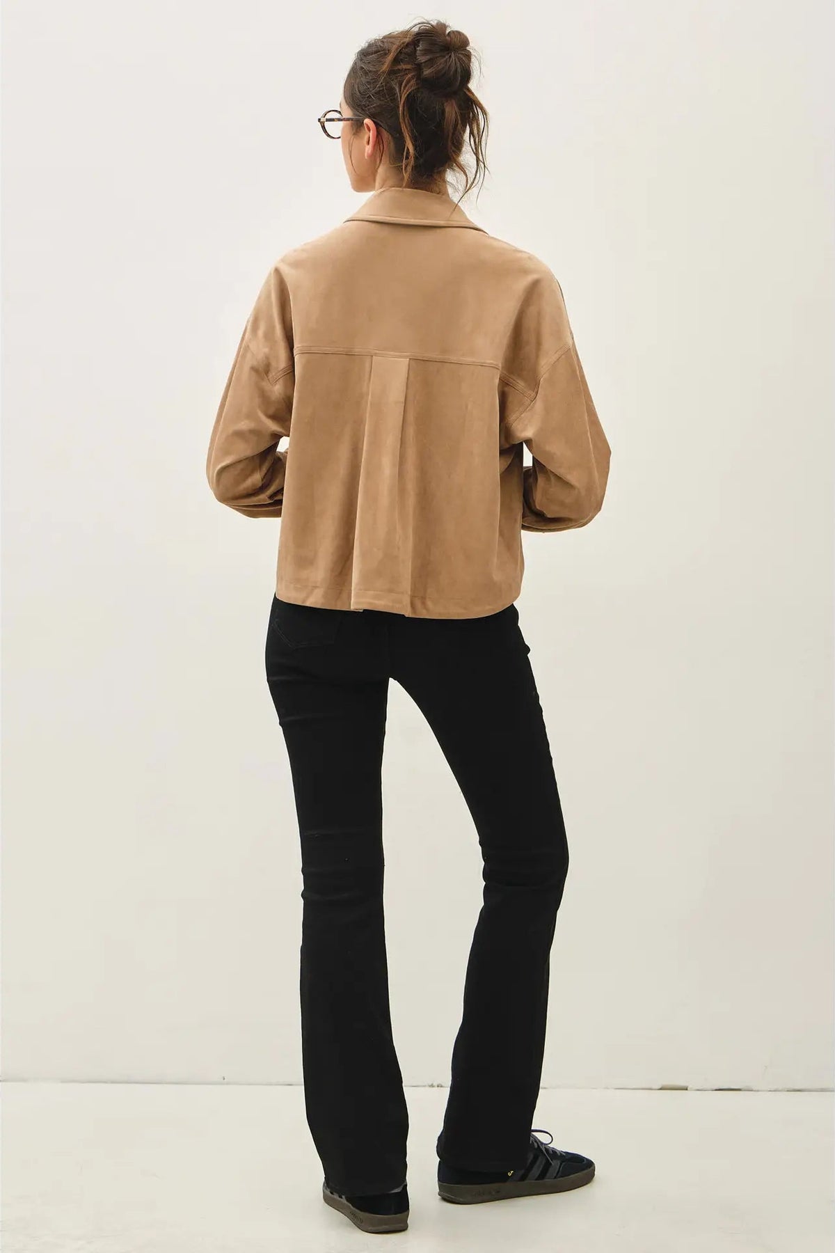 A woman viewed from the back wearing a cropped tan jacket and black pants against a plain white background.