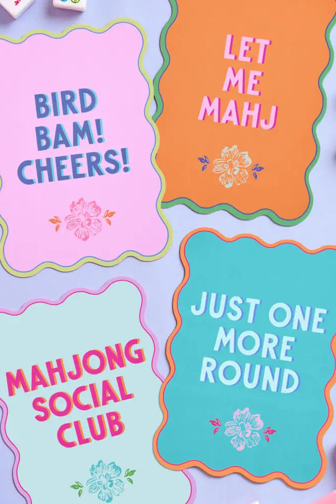 Four colorful wavy-edged cards with Mahjong-themed phrases arranged on a lavender background with scattered white and pink mahjong tiles. The cards read: 'BIRD BAM! CHEERS!' in blue on pink, 'LET ME MAHJONG' in pink on orange, 'JUST ONE MORE ROUND' in white on teal, and 'MAHJONG SOCIAL CLUB' in pink on light blue.