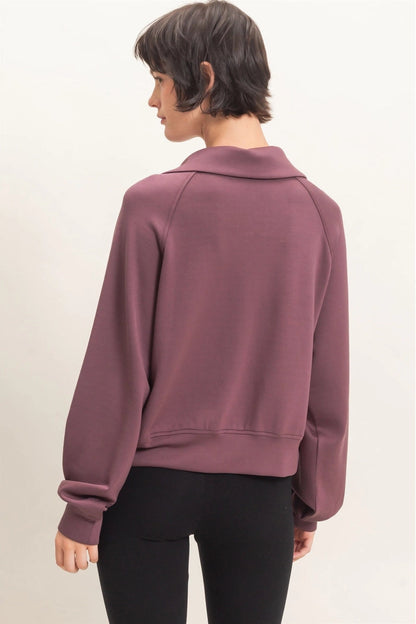 A woman shown from the back wearing a dusty mauve long-sleeve cropped sweatshirt with a mock neck and black leggings, set against a plain off-white studio background.