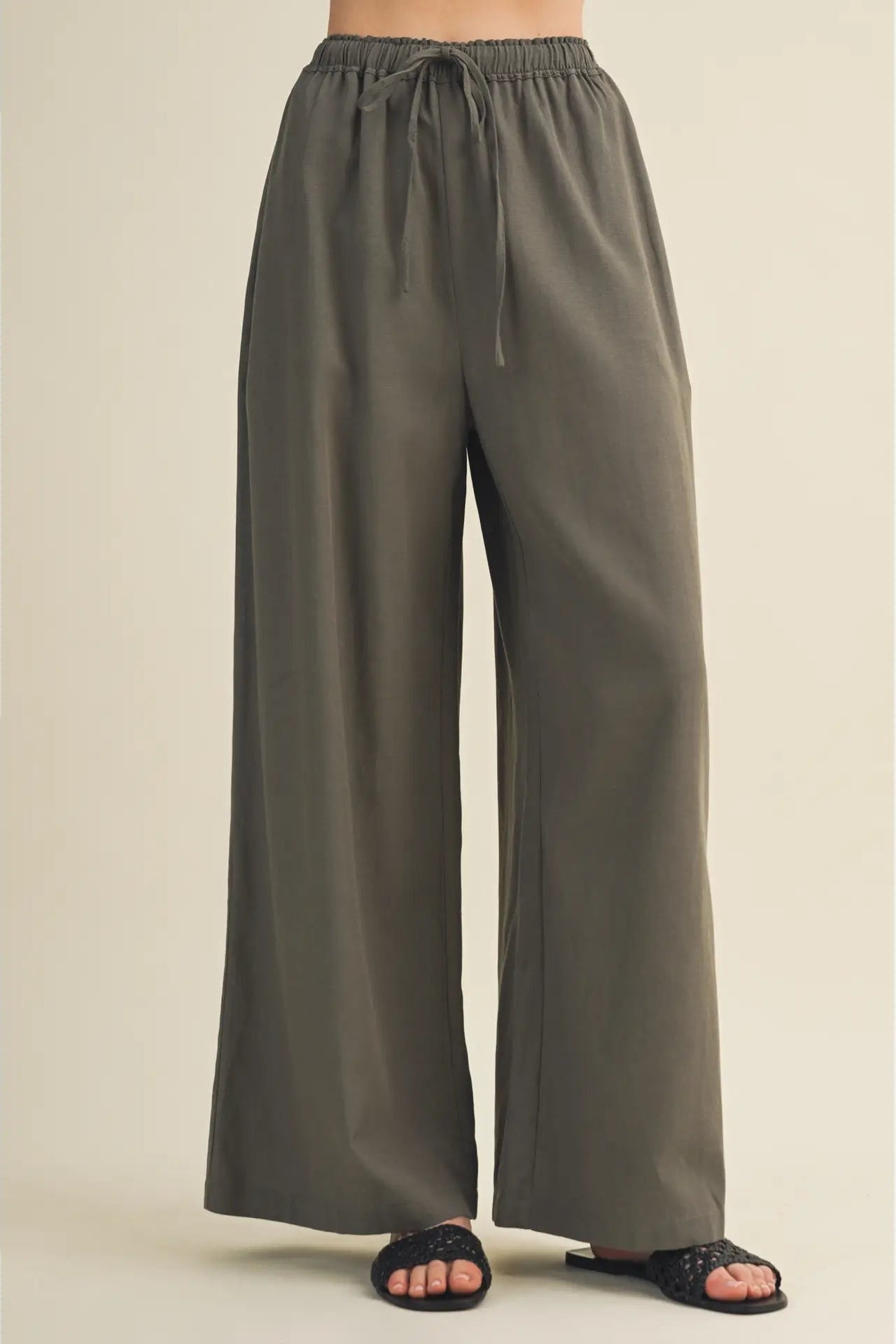 A person wearing wide-leg drawstring pants in a muted olive gray color, paired with black slide sandals, shown against a plain beige studio background.