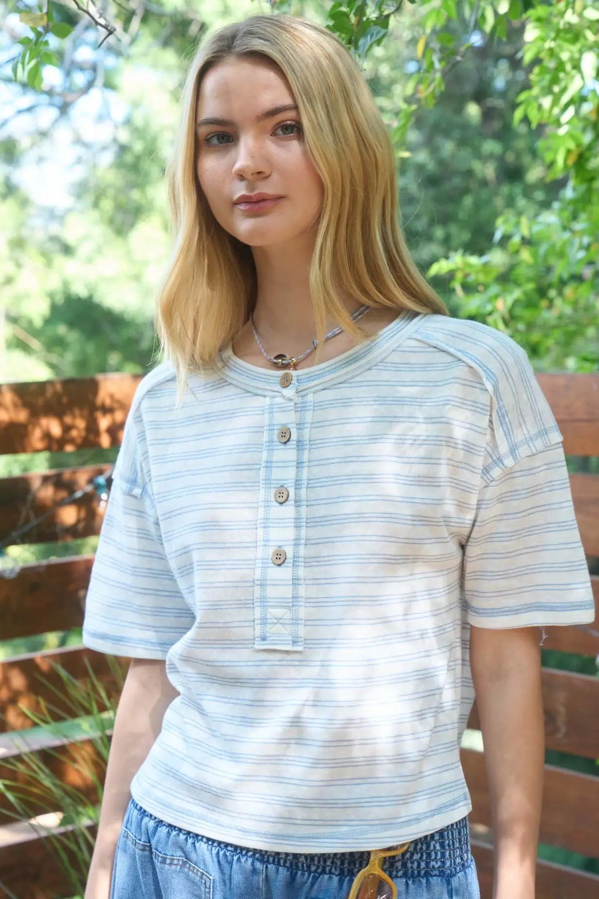 Woman wearing a light blue striped shirt standing outdoors with greenery in the background