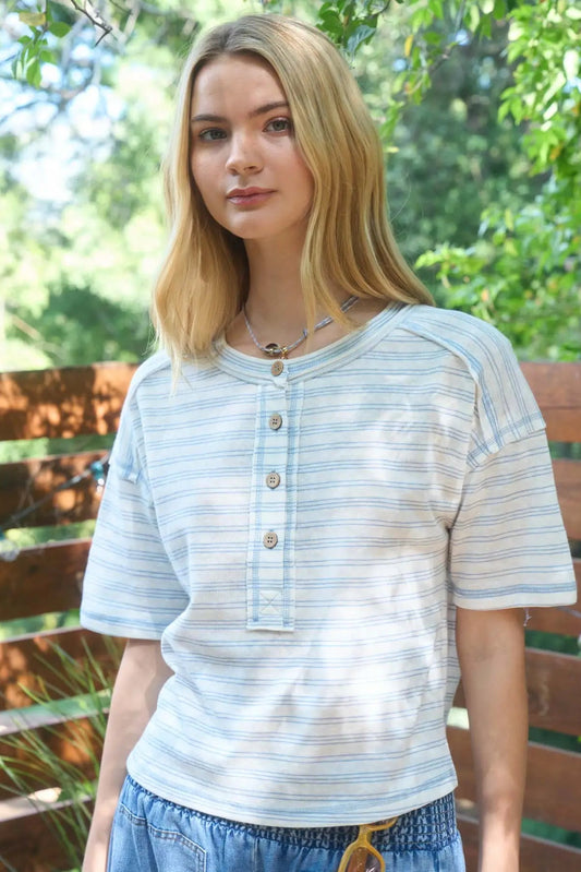 Woman wearing a light blue striped shirt standing outdoors with greenery in the background