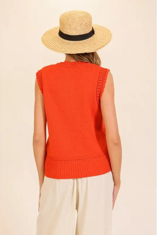 A woman shown from the back wearing a bright orange sleeveless knit vest, a straw hat with a black band, and light beige pants, against a plain cream background.