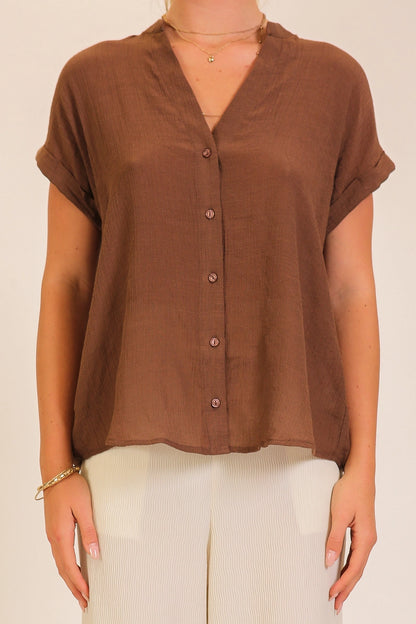 A woman wearing a brown short-sleeve button-up top with a V-neck and white pants, shown from the neck down against a plain light background.