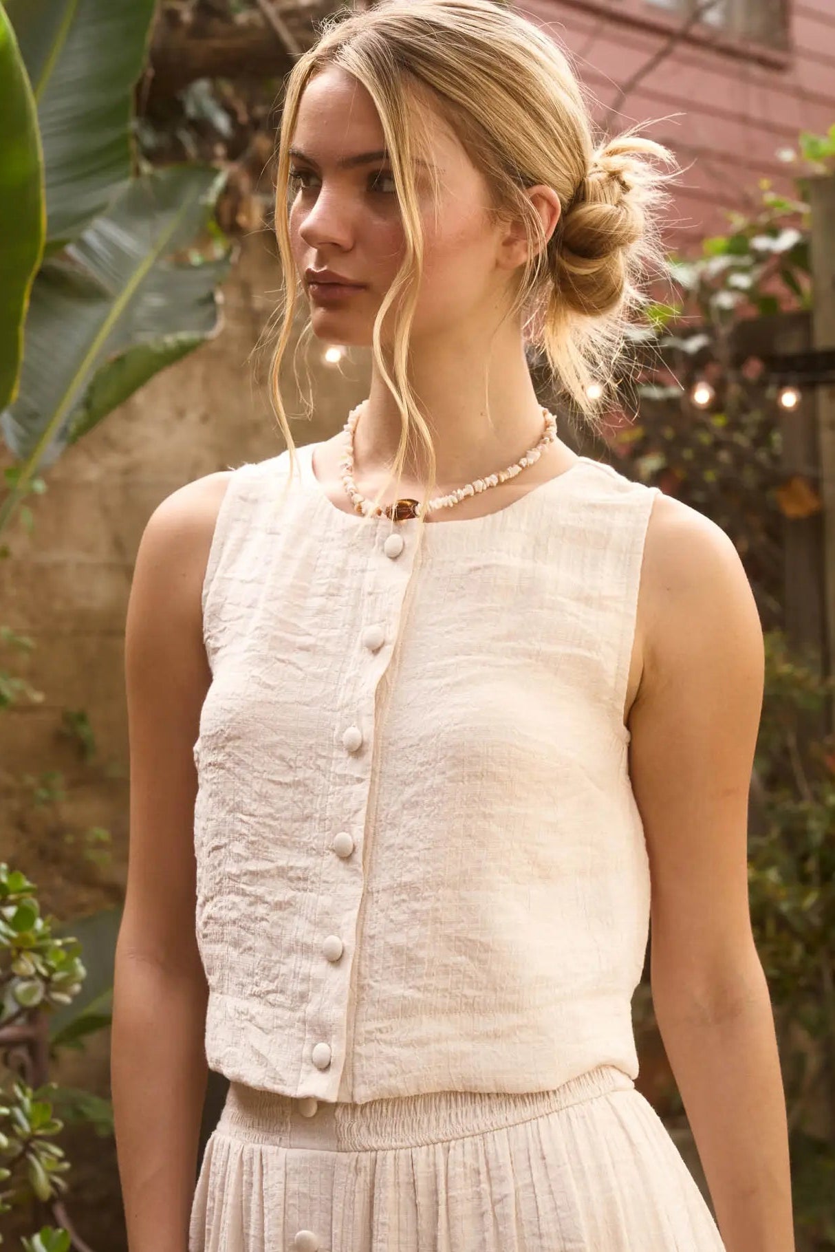 A woman wearing a sleeveless cream-colored textured linen vest with a button front, paired with a matching pleated skirt, set against a warm, garden-like background.