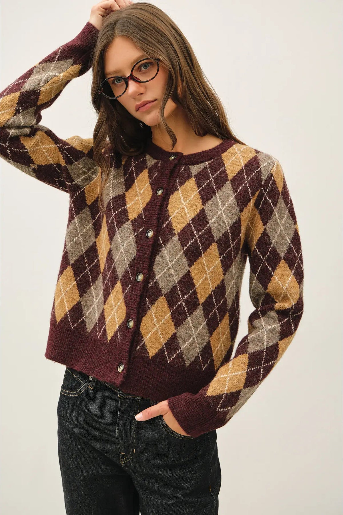 A woman wearing a long-sleeve button-up cardigan with an argyle pattern in burgundy, tan, and beige, paired with dark jeans against a plain light background.
