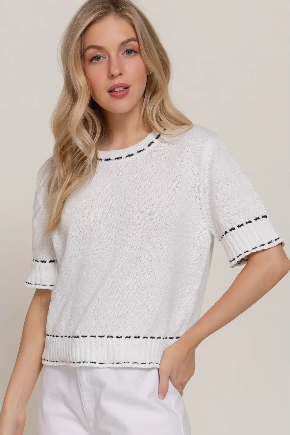 A blonde woman wearing a white short-sleeve knit top with black dashed trim details at the collar, sleeve cuffs, and hem, paired with white pants, set against a plain beige studio background.
