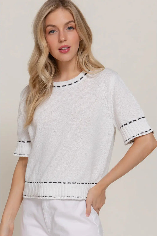 A blonde woman wearing a white short-sleeve knit top with black dashed trim details at the collar, sleeve cuffs, and hem, paired with white pants, set against a plain beige studio background.