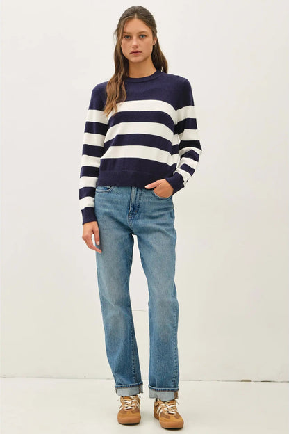 A woman modeling a navy blue and white horizontally striped long-sleeve knit sweater, paired with light-wash blue jeans and brown lace-up shoes, against a plain off-white studio background.
