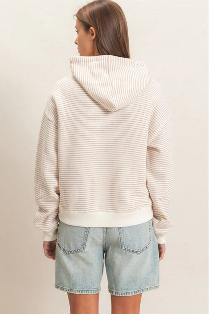 A woman shown from the back wearing a cream-colored striped hooded sweatshirt and light-wash denim shorts against a plain beige studio background.