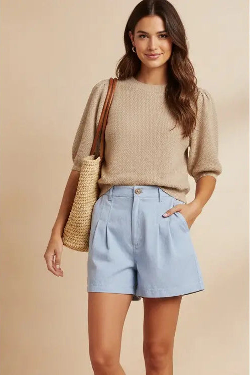 Woman wearing a beige sweater and light blue shorts, holding a straw bag on a beige background