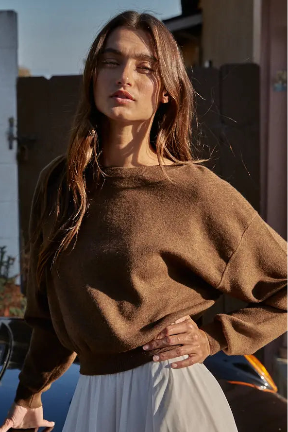 A woman wearing a brown knit crewneck sweater and a white skirt, standing outdoors near a car with a fence and house in the background.
