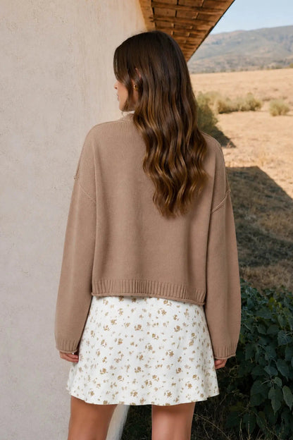 A woman shown from behind wearing a cropped tan knit sweater and a white floral mini skirt, standing outdoors next to a stucco wall with a dry field and hills in the background.