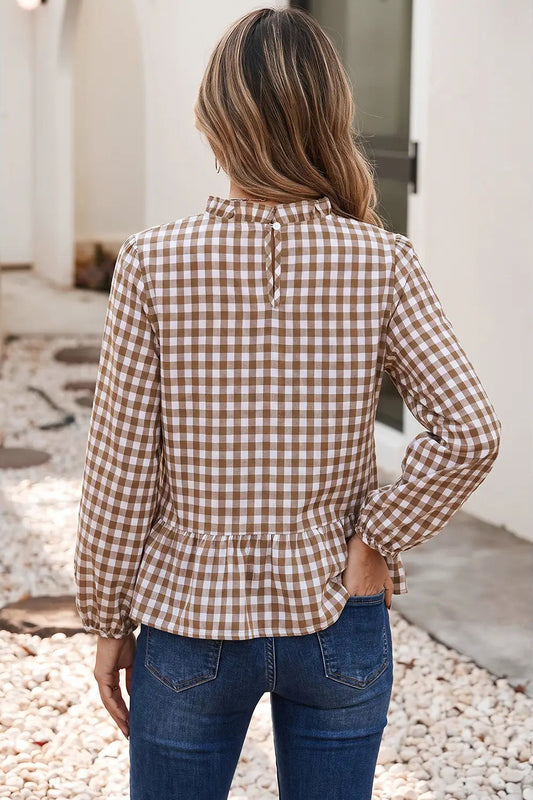 A woman viewed from behind wearing a brown and beige gingham long-sleeve blouse with a ruffled hem, paired with blue jeans, standing outdoors on a stone path.