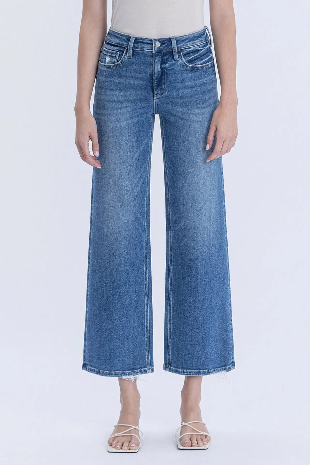 A model wearing a pair of medium-wash blue cropped wide-leg denim jeans with a frayed hem, paired with a light beige top and white strappy sandals, against a plain white studio background.