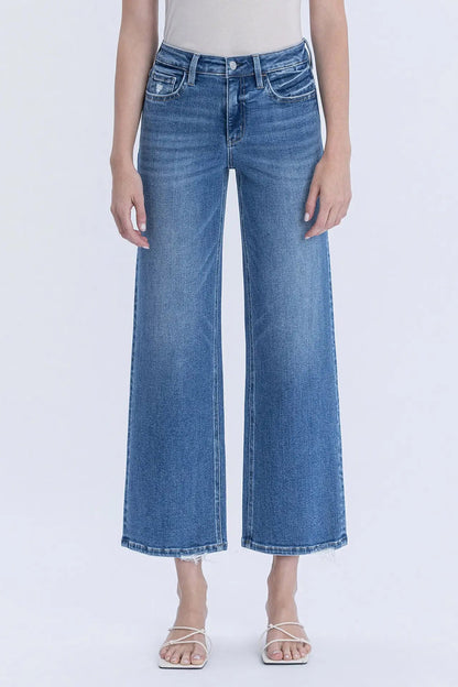 A model wearing a pair of medium-wash blue cropped wide-leg denim jeans with a frayed hem, paired with a light beige top and white strappy sandals, against a plain white studio background.