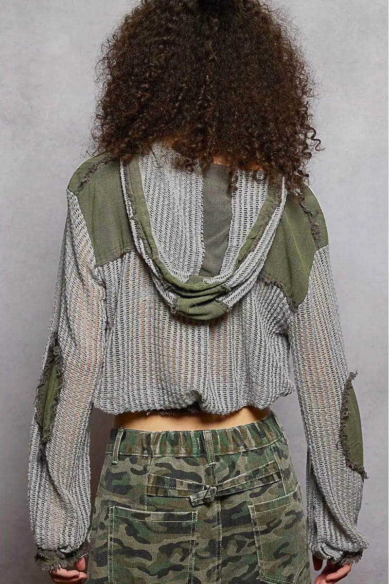 A person shown from the back wearing a cropped, long-sleeve knit top with a striped gray and beige body, olive green shoulder panels, and a draped green neck detail, paired with camouflage pants against a gray studio background.