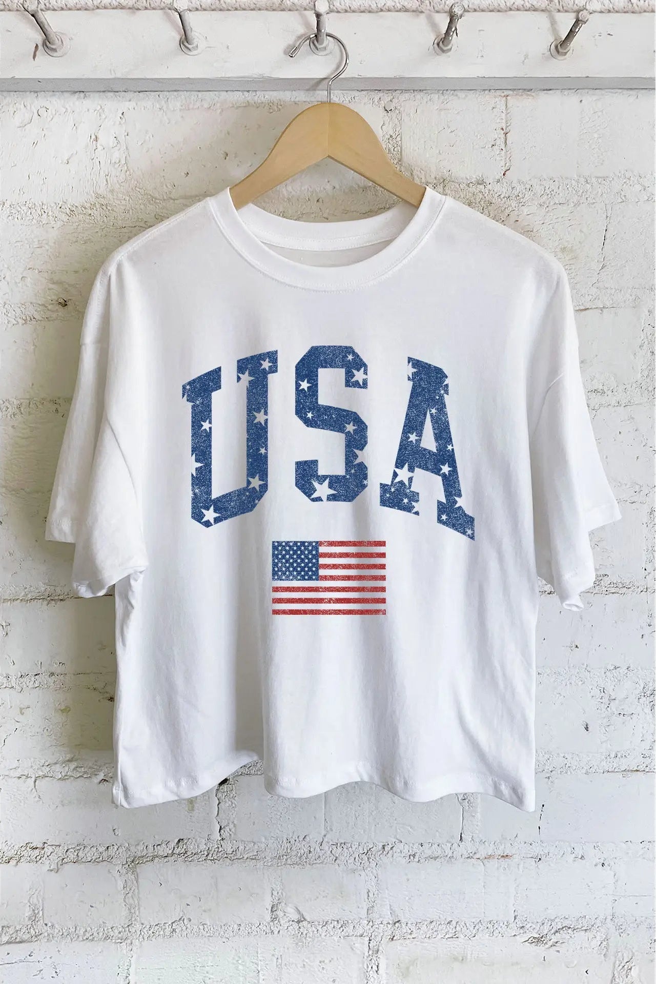 White t-shirt with 'USA' and American flag graphic on a hanger against a white brick wall.