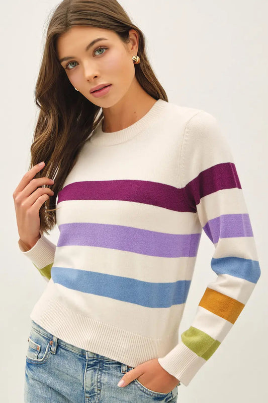 A woman wearing a cream-colored crewneck sweater with horizontal stripes in purple, light purple, blue, yellow, and green, paired with blue jeans, against a plain light background.